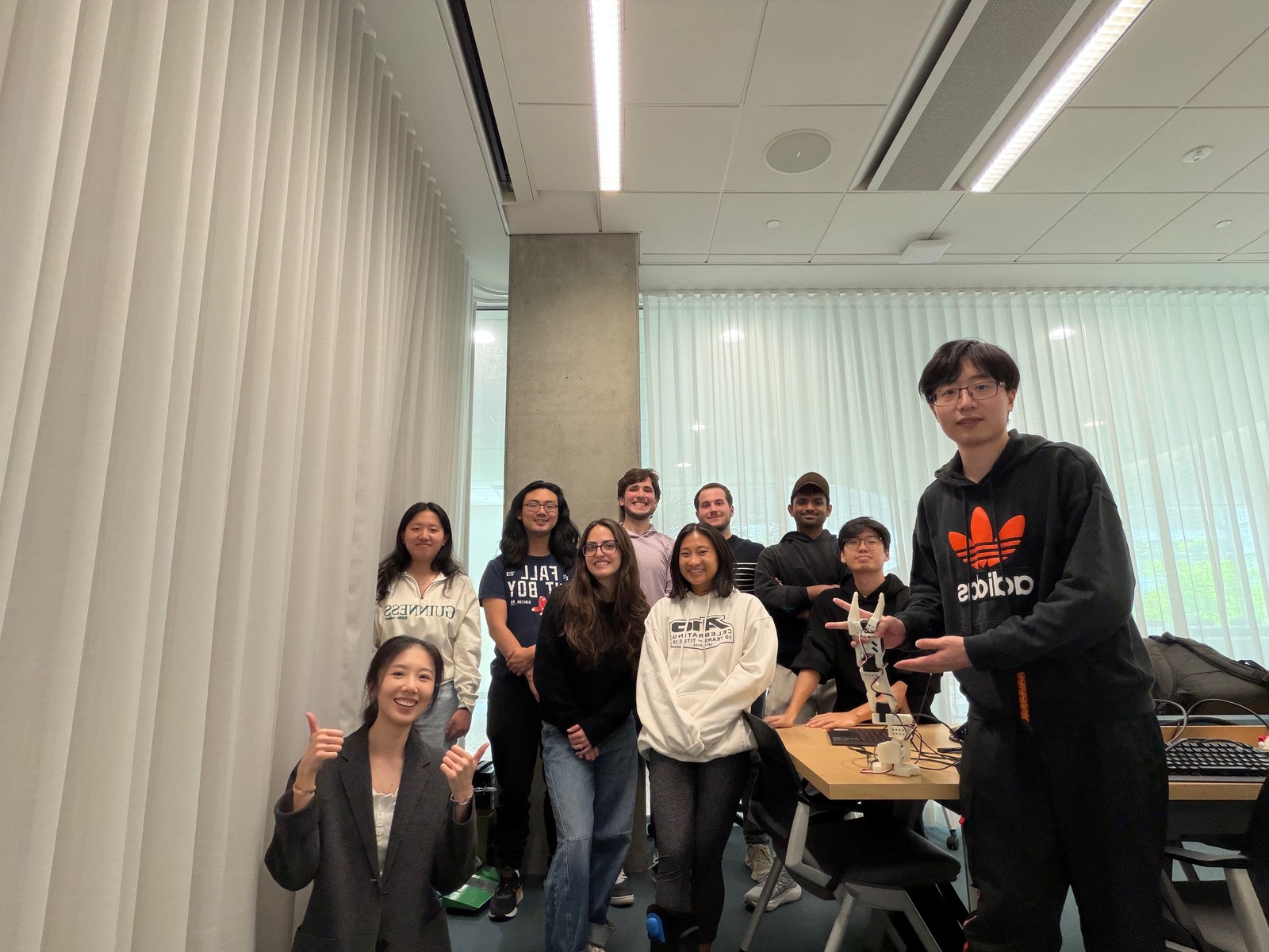 Chuchu Chen giving two thumbs up with her students and a robot arm during their Spring 2026 course-project demonstrations.